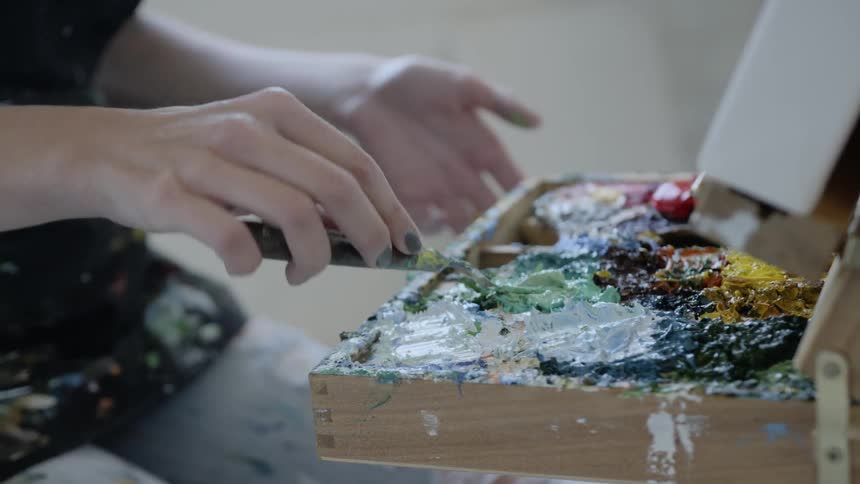 A woman sitting down in front of a canvas while using a utensil to mix paint