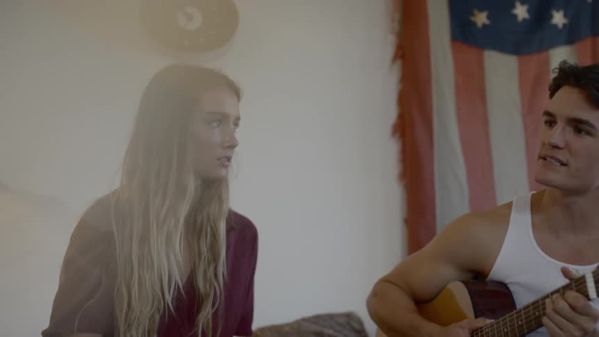 A woman stares at a man sitting beside her holding a guitar and talking