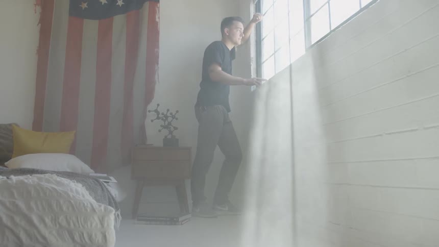 A young man stands and looks out his bedroom window