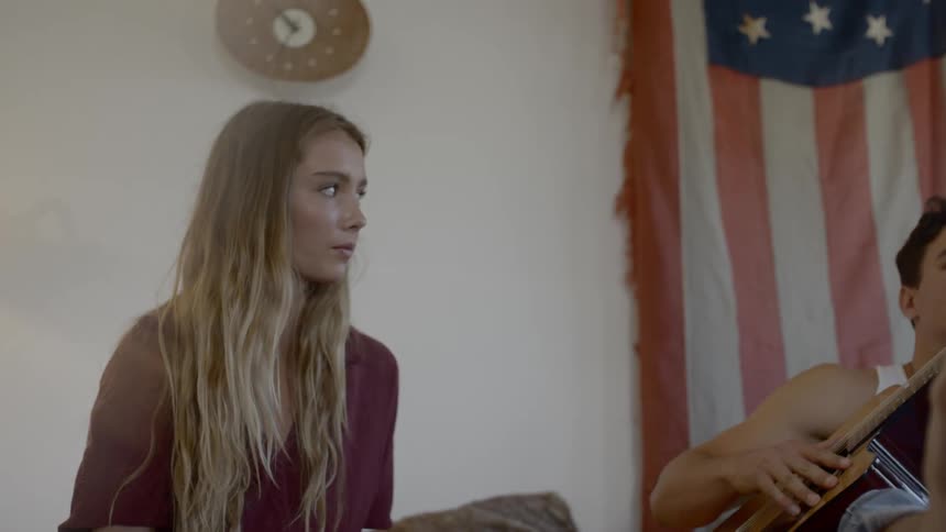 Blonde woman and man who prepares to play the guitar sits on the couch before the american flag in a room