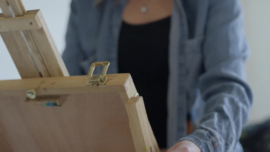 A young woman standing indoors carefully raises a wooden painting easel in front of herself
