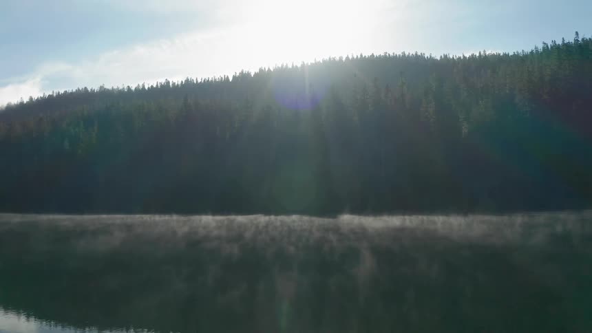The sun rising over a tree lined hill with mist coming off a lake