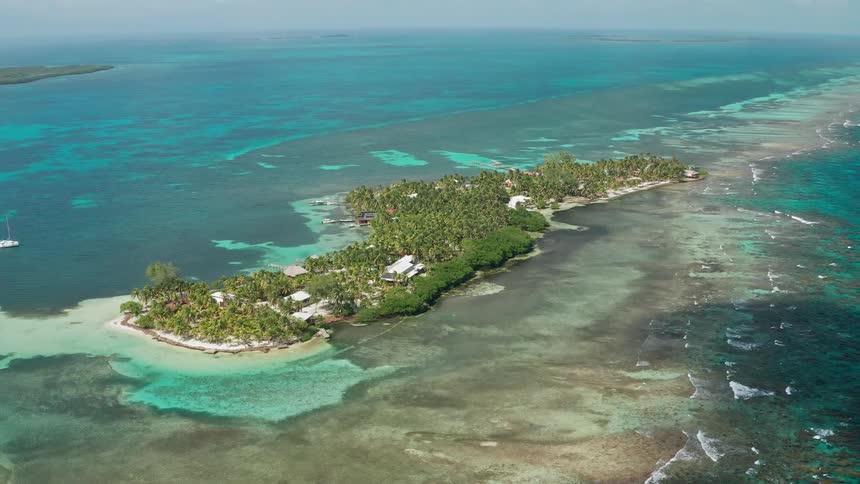A small island full of trees and a few buildings is surrounded sandy shores and ocean water