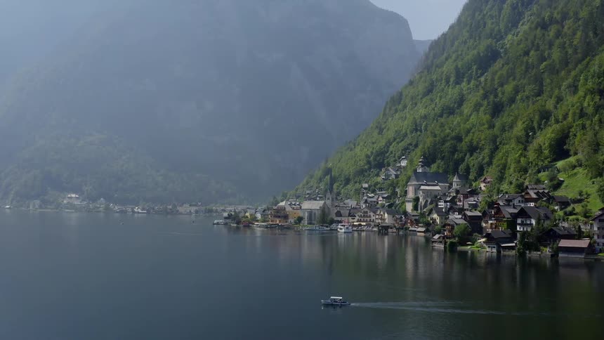 Boat travels across lake next to town at foot of forest covered mountain