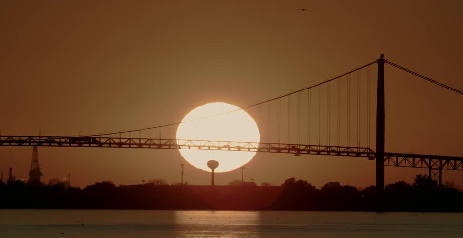 The silhouette of cars and trucks traveling over a ridge with a large glowing sun behind at sunrise