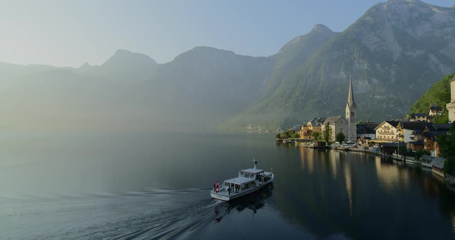 A boat motors slowly toward a dock at the shore of a small village at the base of some mountains on a hazy day