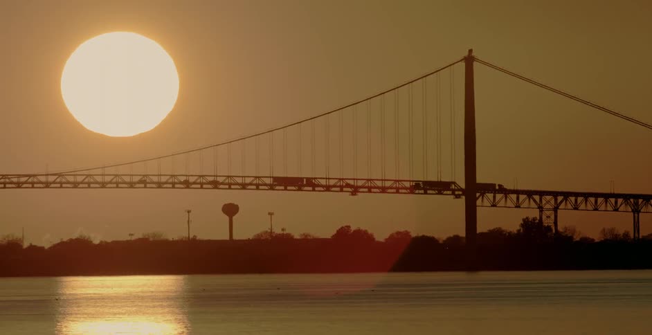 The large glowing sun in the sky behind the silhouette of traffic crossing a bridge