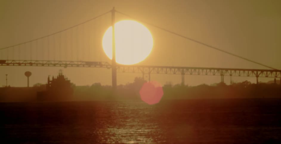 The sun is setting low in the sky behind the silhouette of a suspension bridge over water
