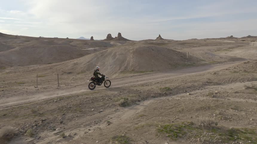 A motorcycle rider drives on a dirt trail in the desert past brush and rock formations