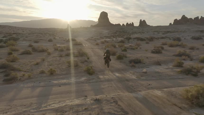 A motorcycle rider drives on a dirt trail past brush and towards rock formations in the desert