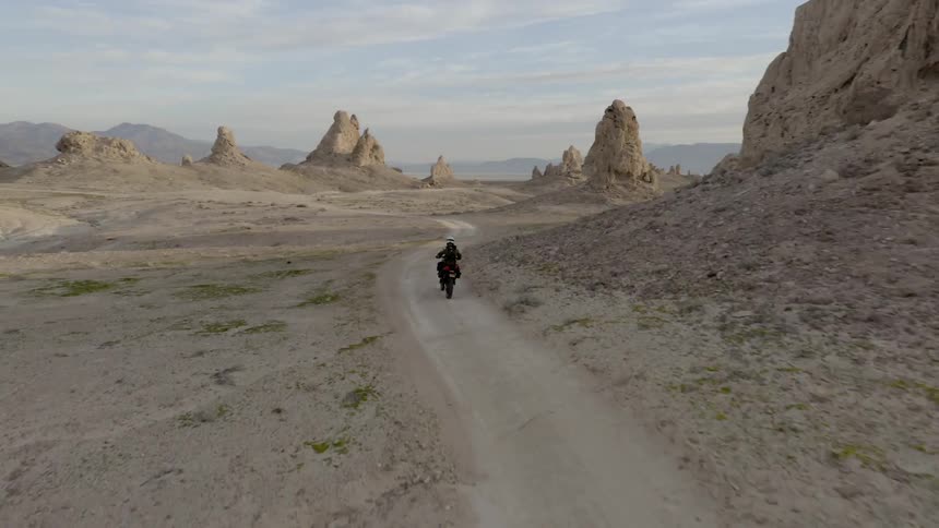 A motorcycle rider drives through the desert on a dirt trail past rock formations