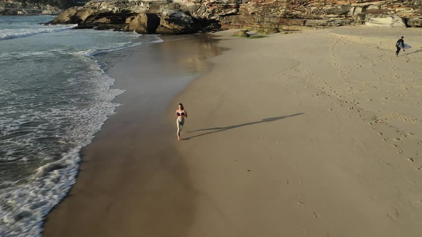 A woman jogs on a sandy beach as waves come to shore and a surfer carries his board away from the water