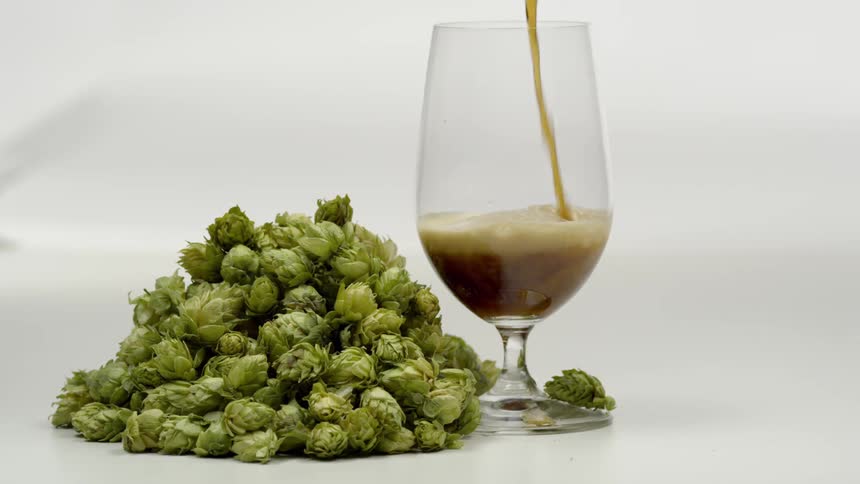 Dark beer is poured into a glass goblet next to small green vegetables