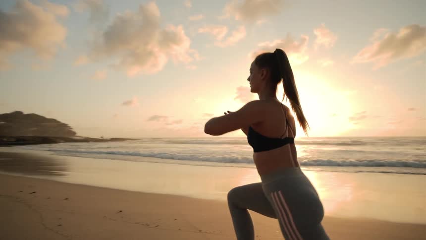 A woman does lunges while exercising on the beach at sunrise