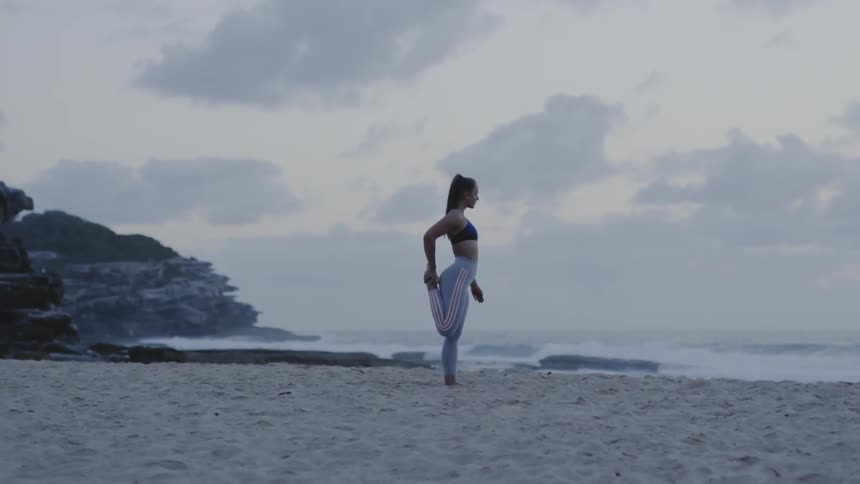 A woman holds her foot behind her as she stretches on the beach sand on a cloudy day