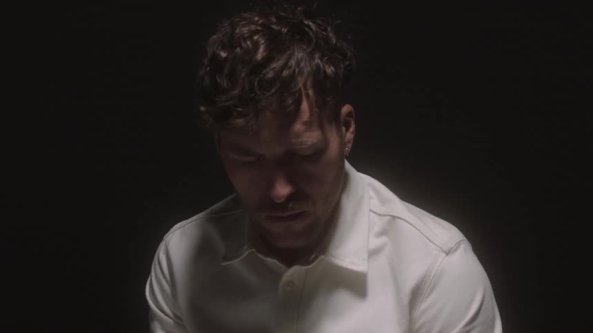 A white man looks down at the ground and turns his head to the side and lights flash behind him