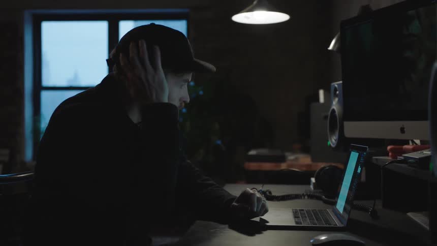 A man sits at his desk and types on a laptop in a dark room