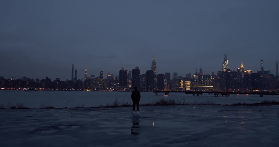 A man stands at the edge of a body of water staring at the city skyline across from him