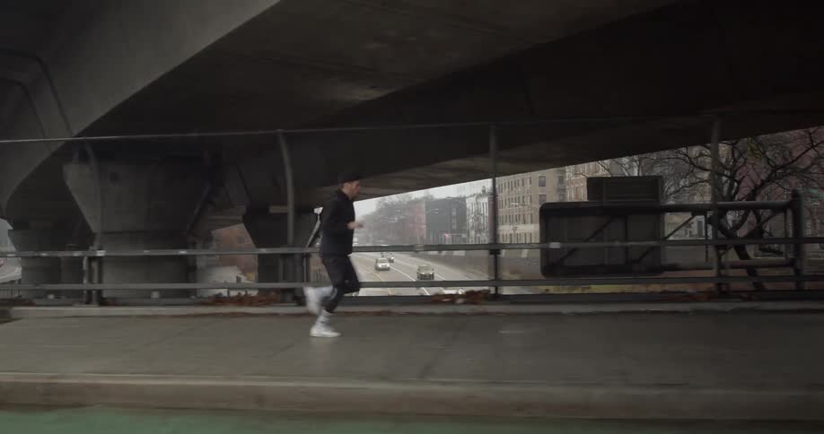 A man jogs on a city sidewalk on an overpass as cars travel by