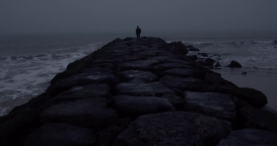 A man stands on a rock pier with waves crashing against the rock