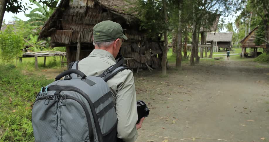 A man in a blue backpack carrying a camera and walking into a village