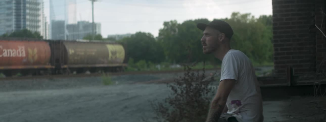 A man sitting down and rubbing his chin with his thumb as he looks into the distance in an urban area near a rail yard