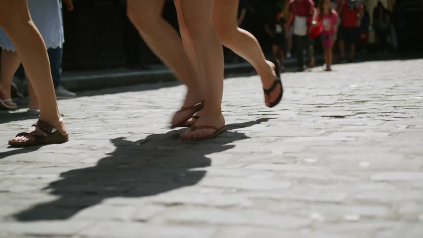 The bare legs of women in flip flops and sandals as they walk over cobble stone on a sunny day