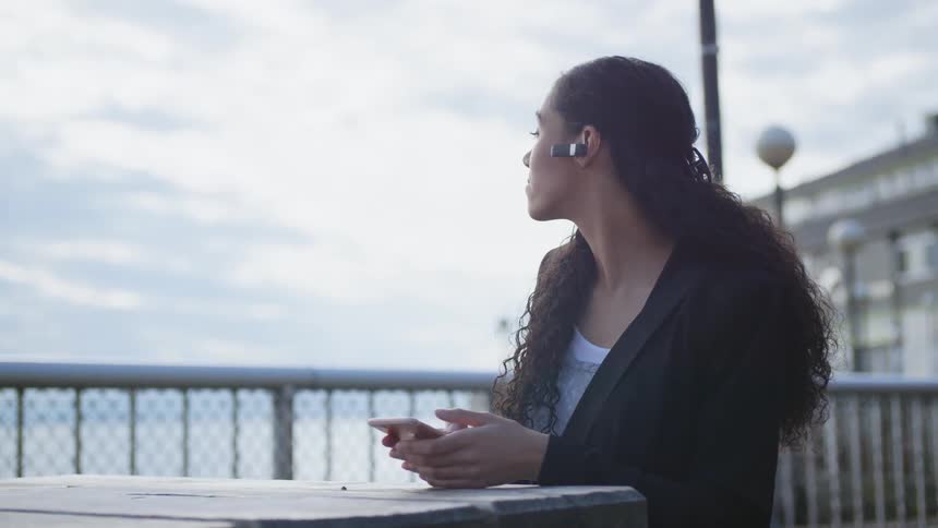 A woman in a formal jacket sitting at a wooden table outdoors wearing an earpiece
