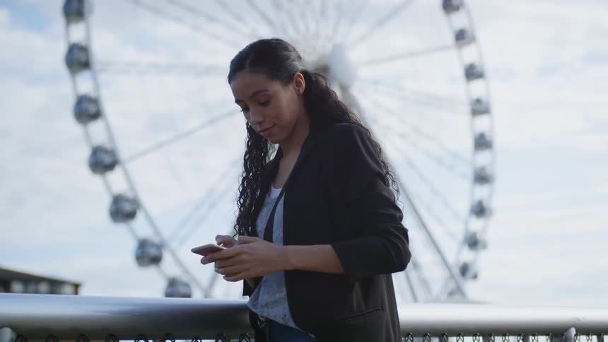 A young woman texting on her phone in front of a ferris wheel at the amusement park