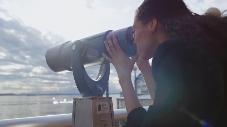 A woman looks through a telescope out towards a body of water