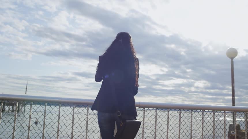 A woman walking towards a public telescope on a pier near the ocean