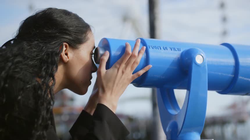 Woman glances through public telescope next to waterfront carnival