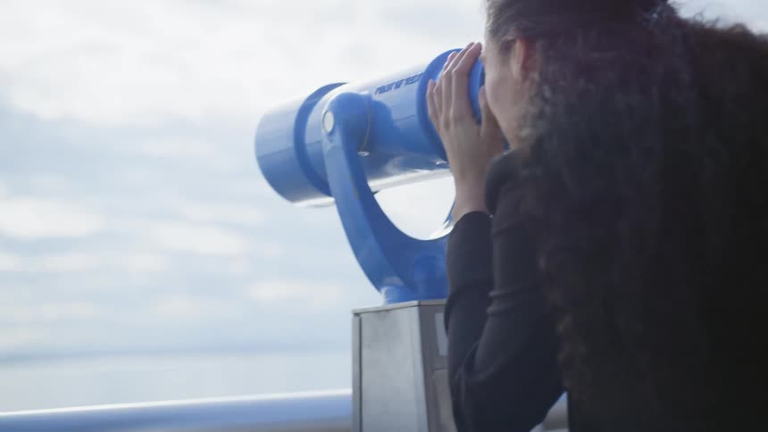 A woman looks through a blue stationary telescope on a pier out to the water