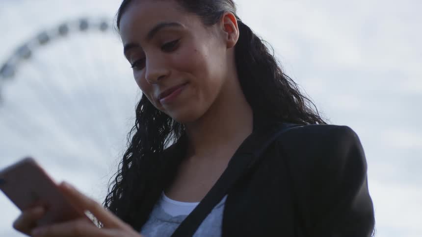 Woman smiles as she views mobile phone near ferris wheel