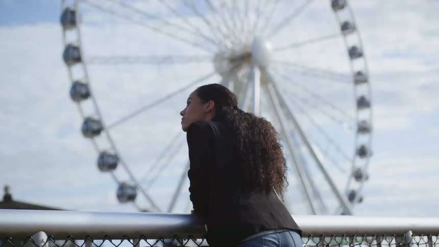 A woman leaning on a fence and looking at the sky as a ferris wheel spins in the background