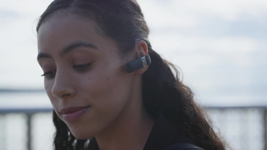 Brunette woman wearing a bluetooth headset looks sideways and holds a smartphone outdoors