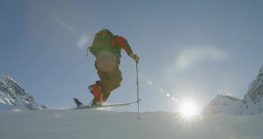 A skier walks on his skis up a hill under a bright sky