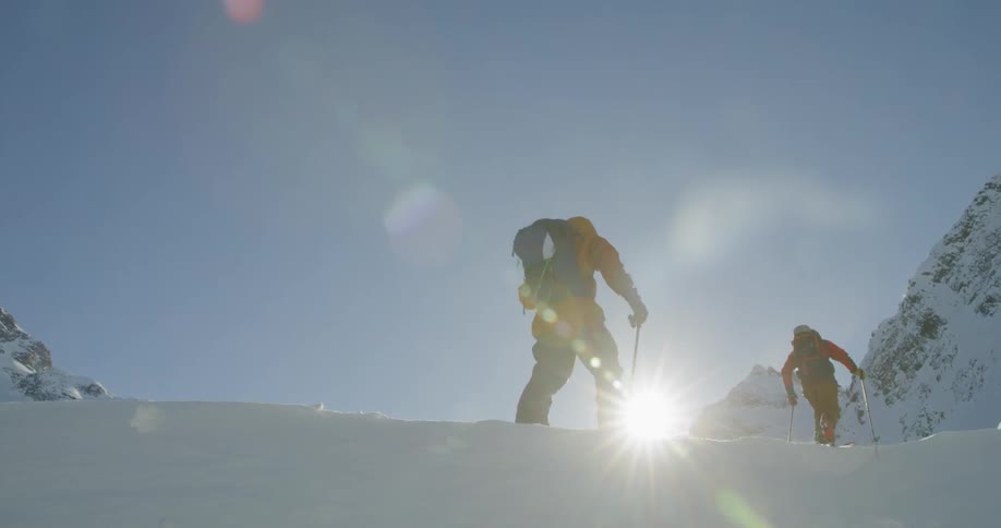 Two people with backpacks on skis walk through the snow towards a mountain on a sunny day