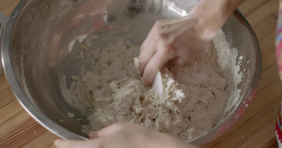 A person mixes dough in a metal bowl on a wood kitchen counter