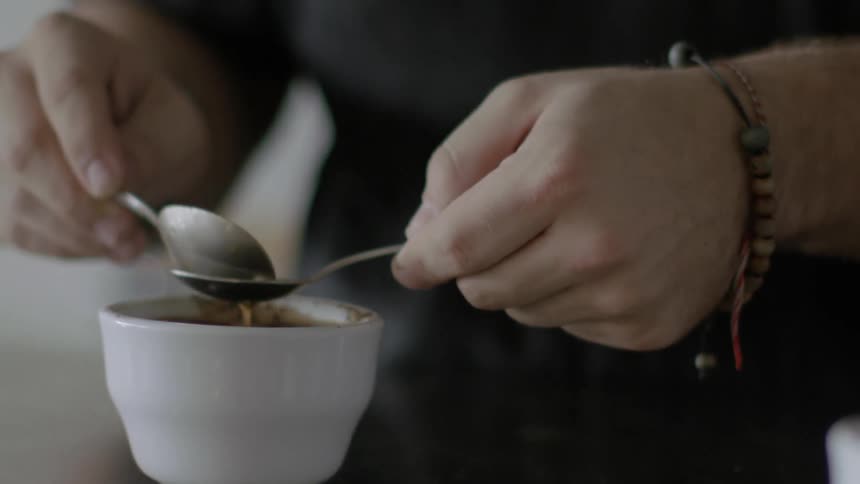 A person carefully uses two small spoons to skim the surface of small cup of coffee before placing it into another nearby cup