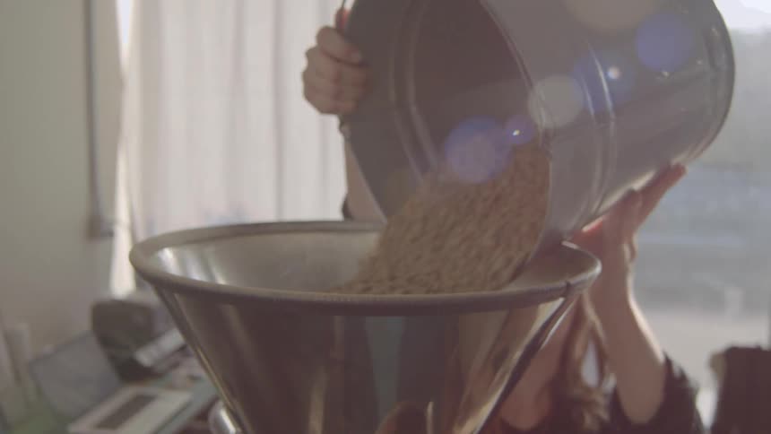 A woman is pouring a large bucket of light colored beans into a large metal funnel atop a roasting machine