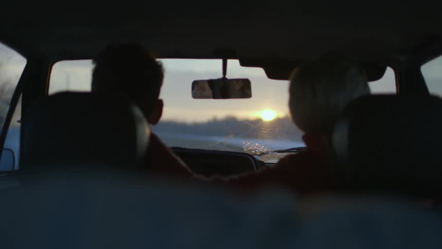 View from the backseat of a woman's hand resting on the back of a man's neck as they drive down a winding, rural road during winter