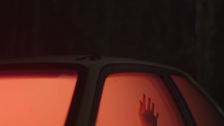A hand is seen rising to touch the window from the interior of a fog-filled car with an orange glow
