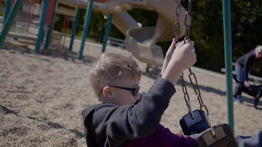 A young boy wearing sunglasses playfully swings on a swing set at an outdoor park