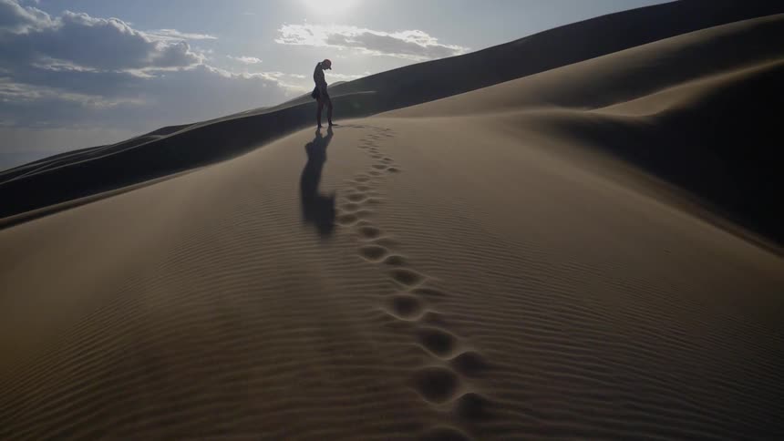 The shadow of person standing atop a sand dune as their body casts a shadow on their footprints below