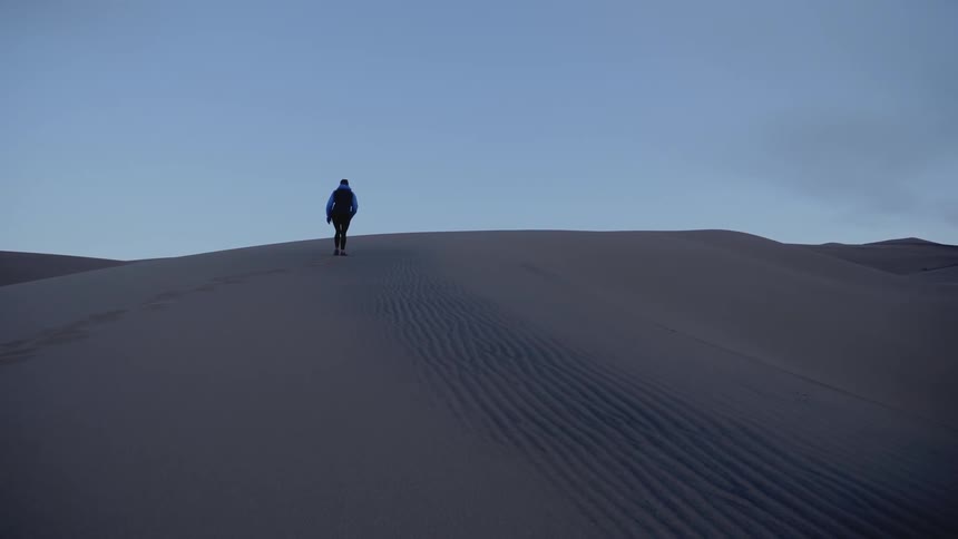 A person climbs to the top of a sand dune leaving a line of footprints near ripples