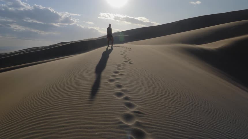 A woman stands at the top of a sand dune with her footprints leading up along the rippling sand