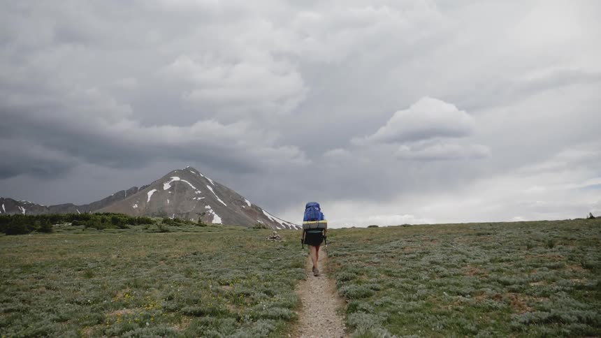 Hiker wearing a backpack walks along a path in the field on a cloudy day