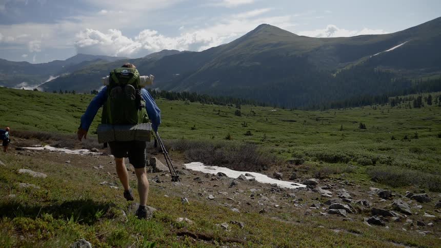 A hiker runs across a grassy field with a mountain range in the distance