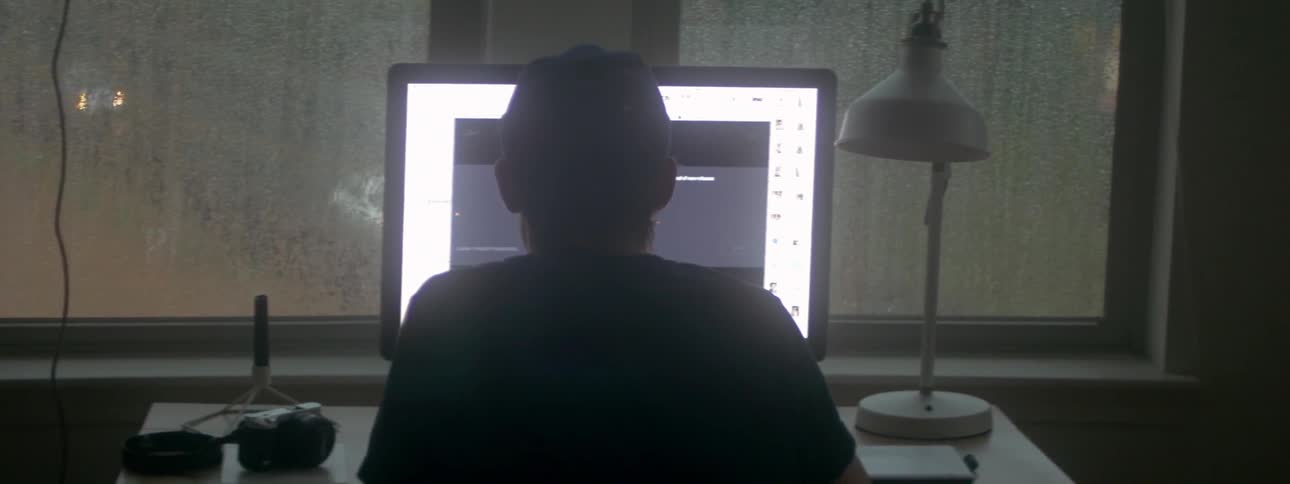 A view from behind of a man using a computer and a drawing tablet in the evening
